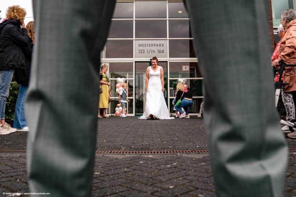 Beeld door de benen van de bruidegom waarop de bruid verguld naar de rug van de bruidegom kijkt voor de first look op hun bruiloft in Rijnsburg © Stefan van Beek Fotografie