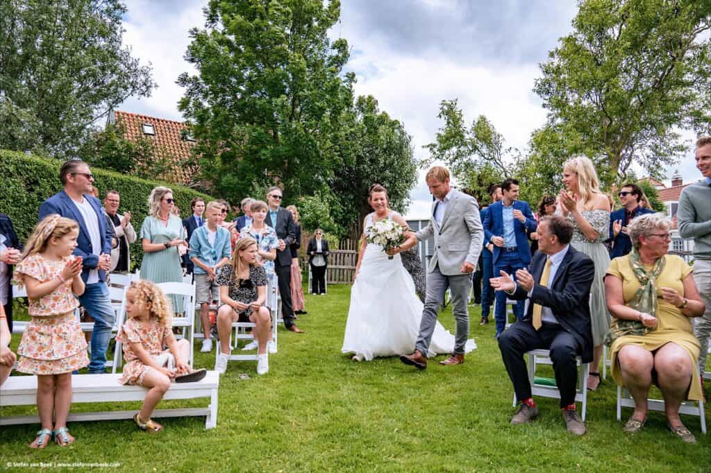 Bruid komt met broer tussen de gasten door aangelopen tijdens start ceremonie bruiloft bij trouwlocatie De Meerhoeve in Oud Ade © Stefan van Beek Fotografie