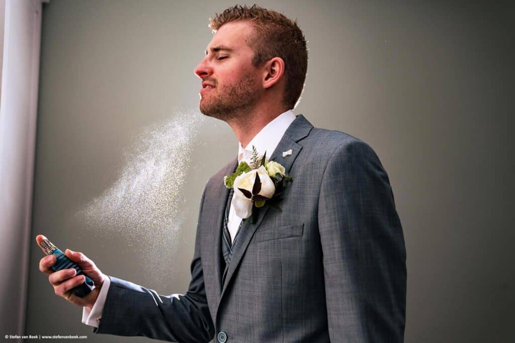 Bruidegom staat met ogen dicht en spayt parfum op met zijn rechter hand waarbij de parfum druppels zichtbaar zijn © Stefan van Beek Fotografie