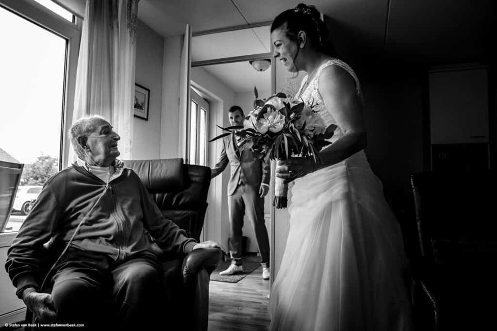 Bruidspaar brengt op hun trouwdag een bezoek aan opa die in een stoel voor het raam zit bij hem thuis © Stefan van Beek Fotografie
