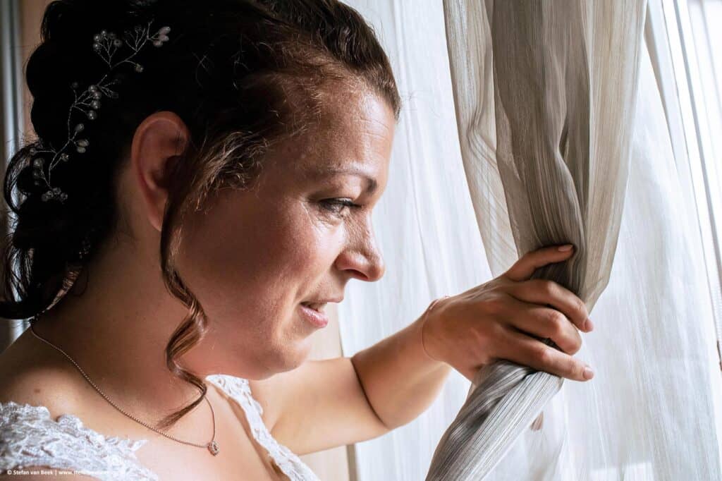 Close-up gezicht en hand van de bruid voor een raam terwijl ze met linkerhand het gordijn openhoud en naar buiten kijkt © Stefan van Beek Fotografie