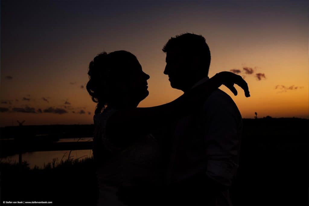 Silhouette van het bruidspaar tijdens creatieve bruidsfotoshoot bij zonsondergang met knaloranje lucht trouwlocatie de Meerhoeve in Oud Ade © Stefan van Beek Fotografie