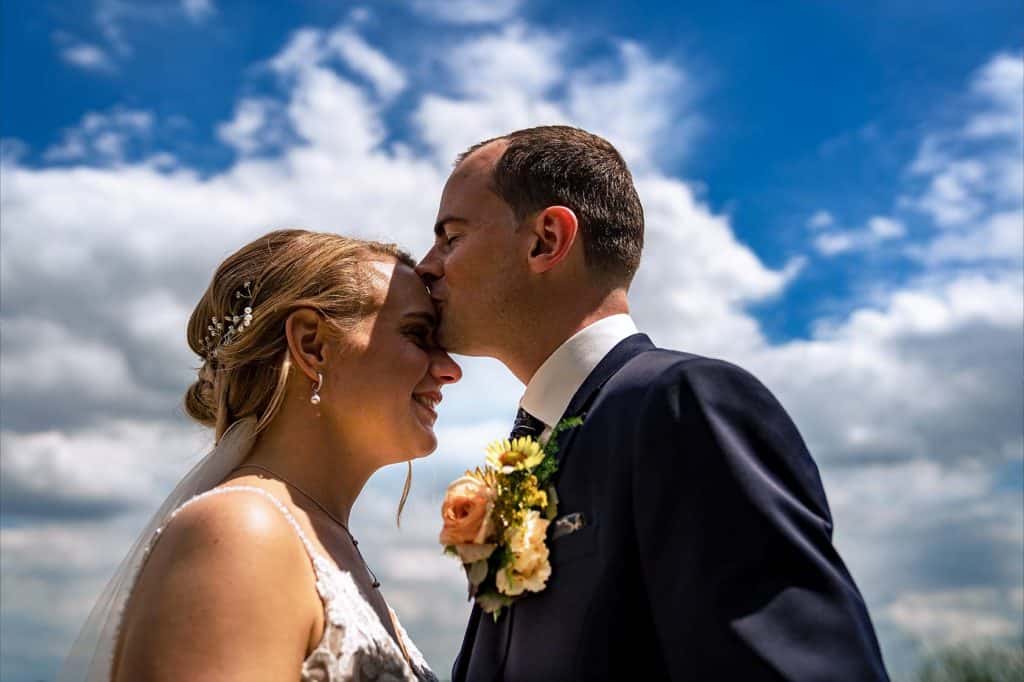 Bruidegom geeft bruid zoen op haar voorhoofd met op de achtergrond de blauwe lucht en wolken © Stefan van Beek Fotografie