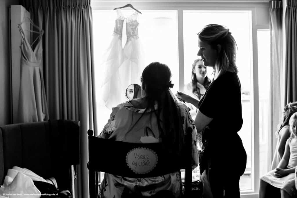 Bruid zit in make-up stoel in hotelkamer Calton Beach Scheveningen en kijkt in spiegel met jurkjes die voor het raam hangen © Stefan van Beek Fotografie