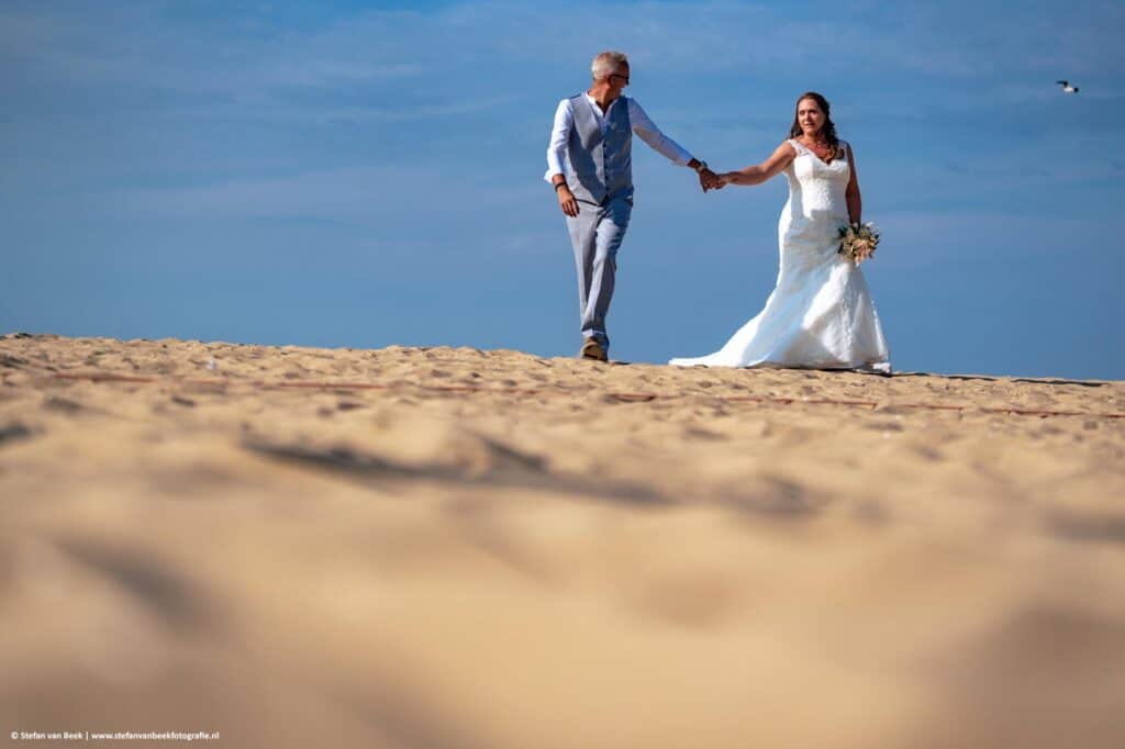 Bruidspaar wandelt hand in hand op het strand tijdens bruiloft op het strand van Scheveningen © Stefan van Beek Fotografie