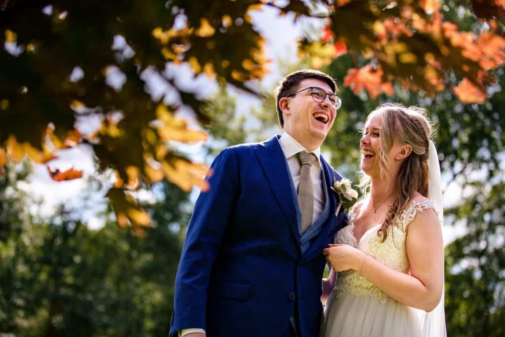 Bride and groom laughing while standing side by side between autumn leaves © Stefan van Beek Photography