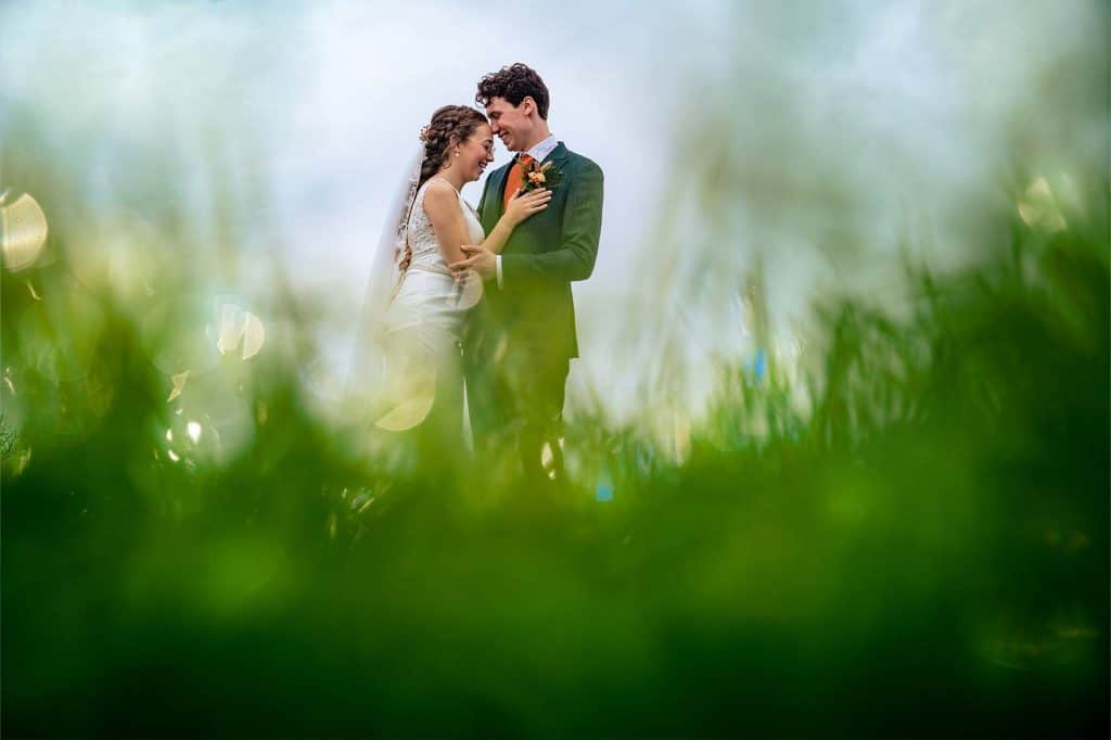 Bride of wedding couple leans while laughing head towards groom which softly touches her head with his nose in between of the grass with bokeh © Stefan van Beek