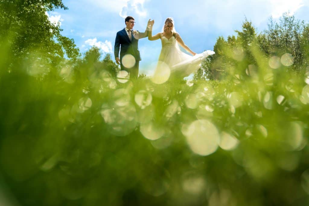Bruidegom laat bruid met haar hand draaien op een groene grasheuvel tegen een blauwe achtergrond tijdens bruiloft fotoshoot © Trouwfotograaf Stefan van Beek