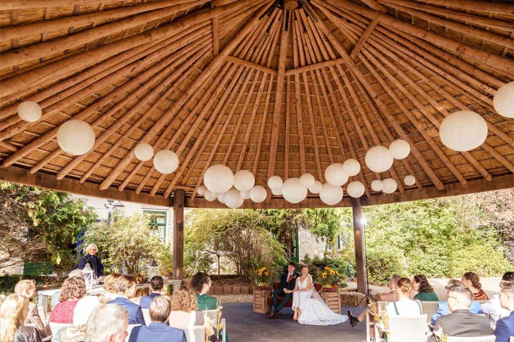 Bruidsceremonie bij trouwlocatie het Wapen van Zoetermeer onder de hooibarg met hout en rieten dak en hangende witte lampionnen © Stefan van Beek