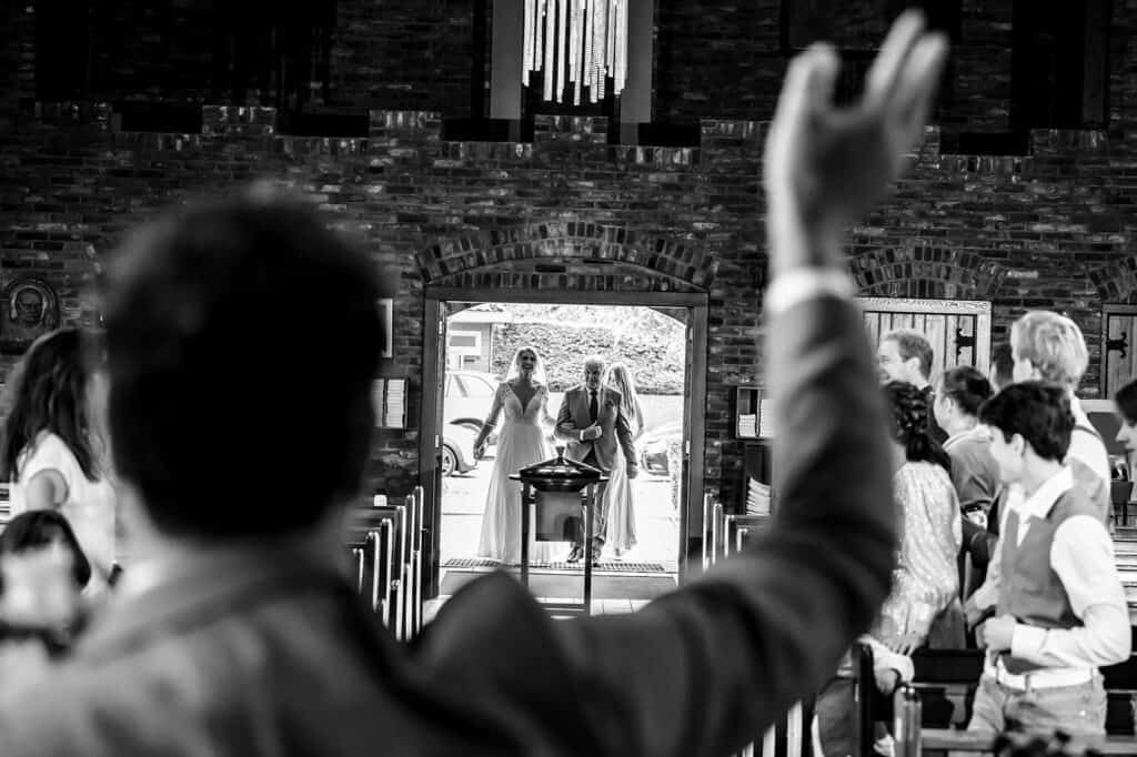 Bruidegom zwaait naar bruid die arm in arm met haar vader de Onbevlekt Hart van Maria kapel in Den Haag in loopt door het gangpad © Stefan van Beek Fotografie