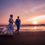 Bruidspaar loopt tijdens zonsondergang met betoverende paars oranje gele lucht hand in hand over het strand terwijl zon nog net boven de zee zichtbaar is © Stefan van Beek Fotografie