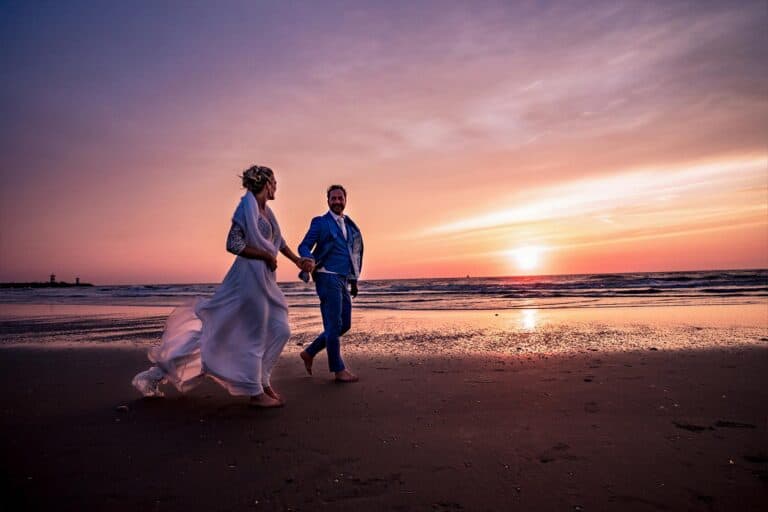 Bruidspaar loopt tijdens zonsondergang met betoverende paars oranje gele lucht hand in hand over het strand terwijl zon nog net boven de zee zichtbaar is © Stefan van Beek Fotografie